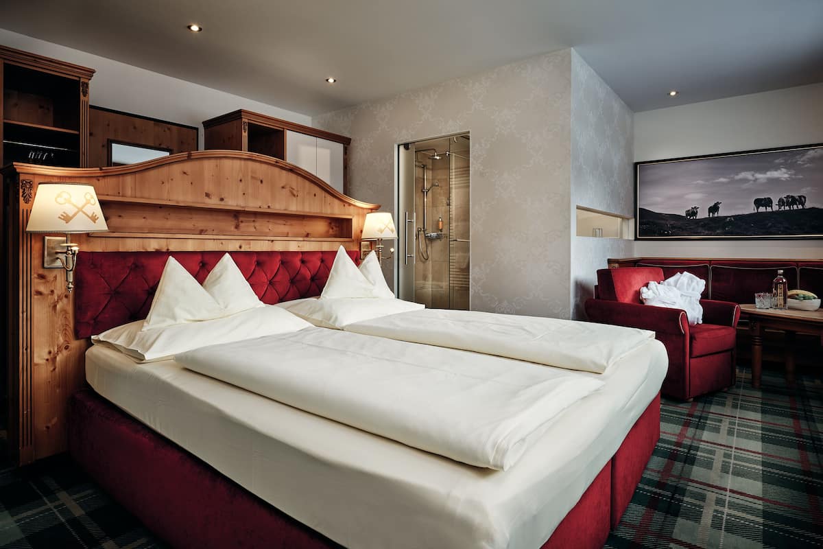 A hotel room with a large bed, a wooden headboard, a red armchair, a desk with a chair, and a large framed picture on the wall.