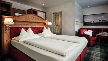 A hotel room with a large bed, a wooden headboard, a red armchair, a desk with a chair, and a large framed picture on the wall.