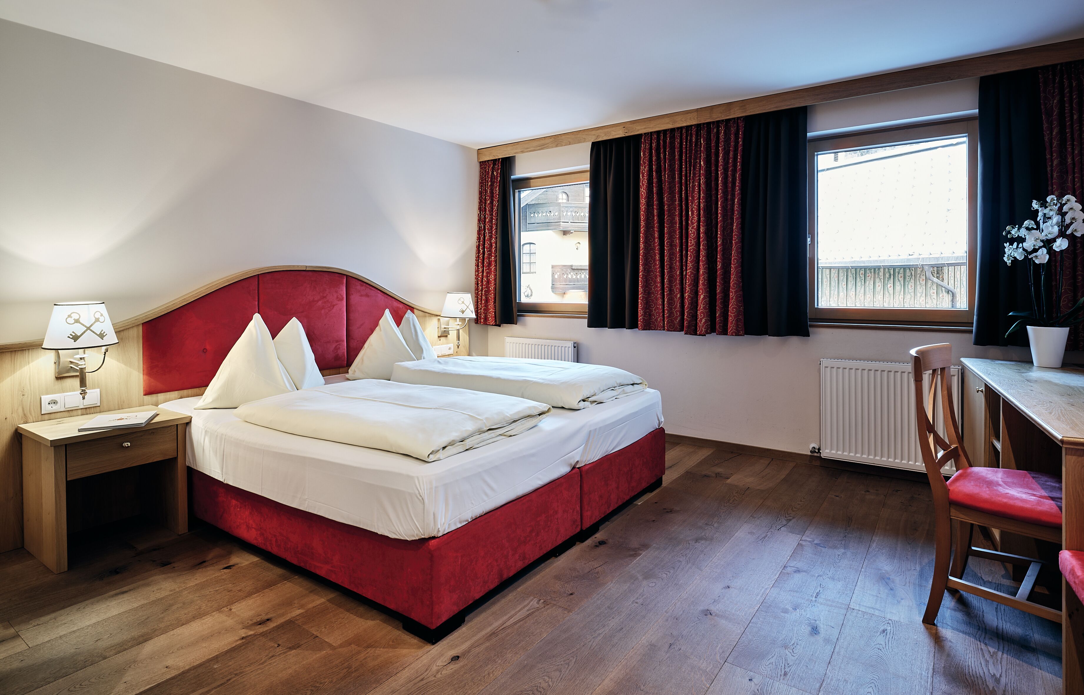 Ein Hotelzimmer mit einem rot gepolsterten Bett, zwei Nachttischen mit Lampen, einem Holztisch mit Stuhl und einem Fenster mit Vorhängen.