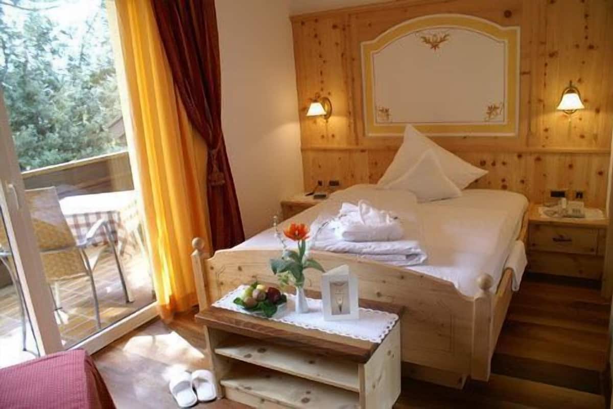 A wooden bedroom with a bed, two bedside lamps, a nightstand, a window with curtains, and a small table with a flower and a bottle.