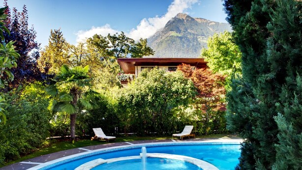 A swimming pool with a fountain, surrounded by lush greenery and a mountain in the background. - Sittnerhof (Merano)
