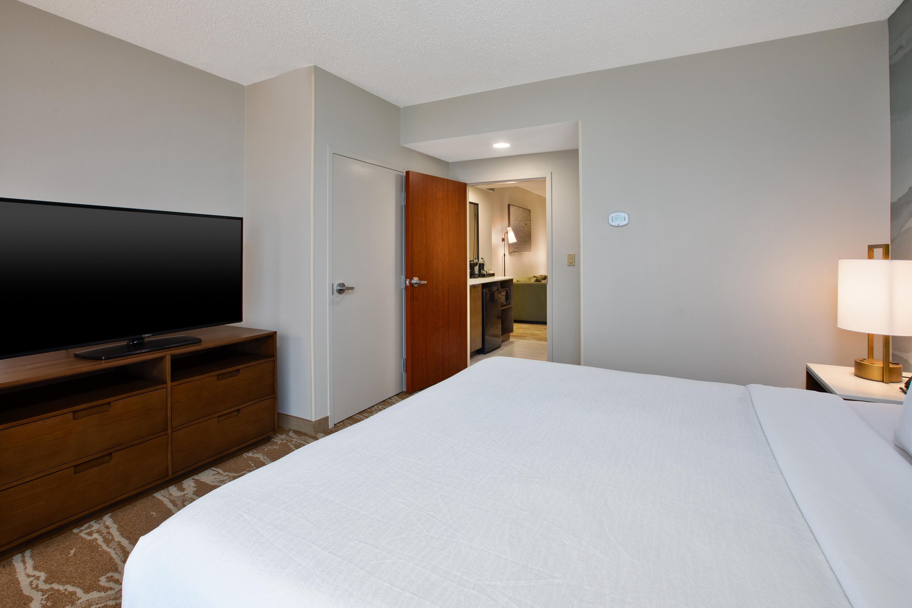 Una habitación de hotel con una cama grande, un televisor de pantalla plana sobre un aparador de madera, una mesita de noche con una lámpara y un baño visible a través de una puerta abierta.