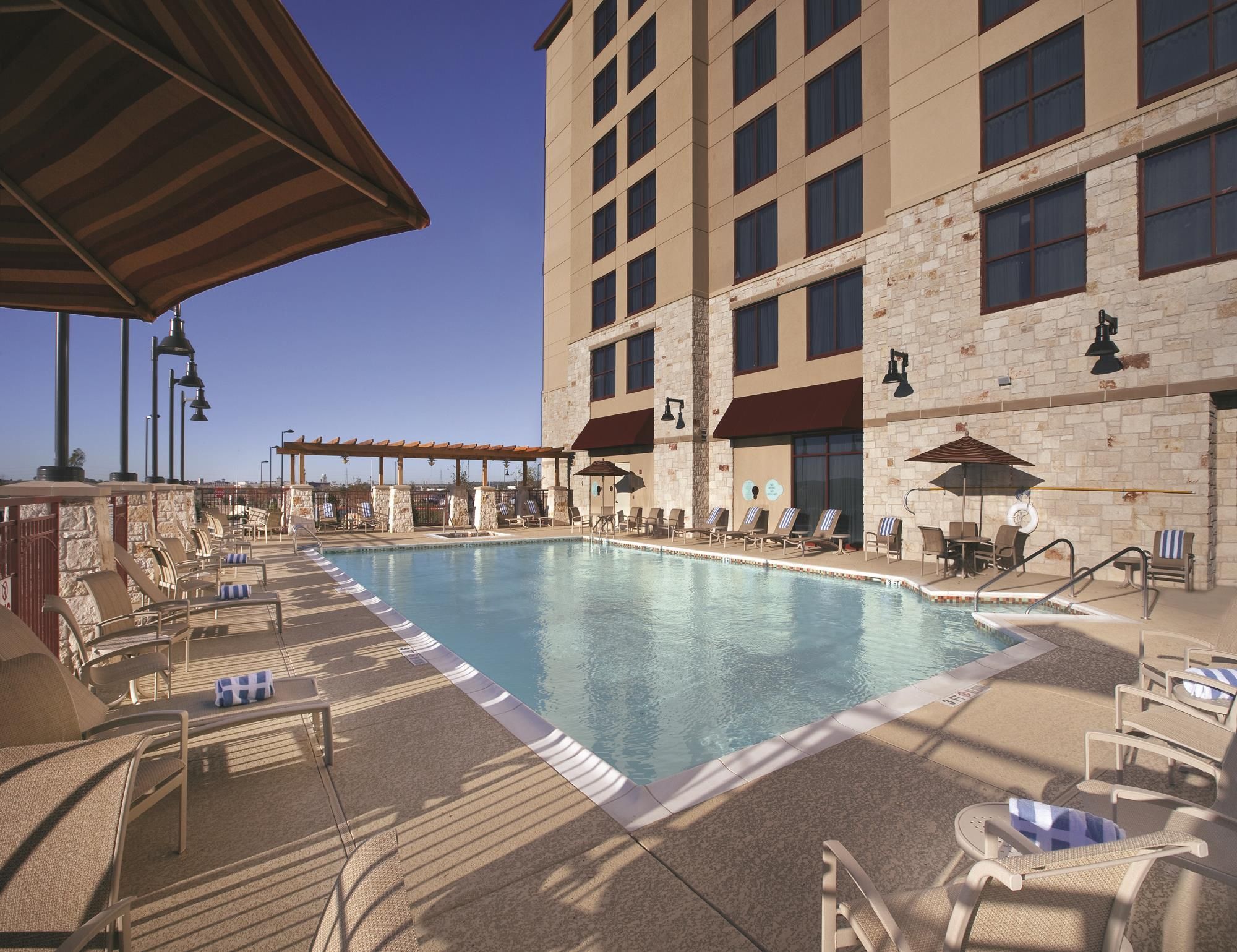 A hotel pool area with lounge chairs and umbrellas, a clear sky, and a building with multiple windows and a stone facade.