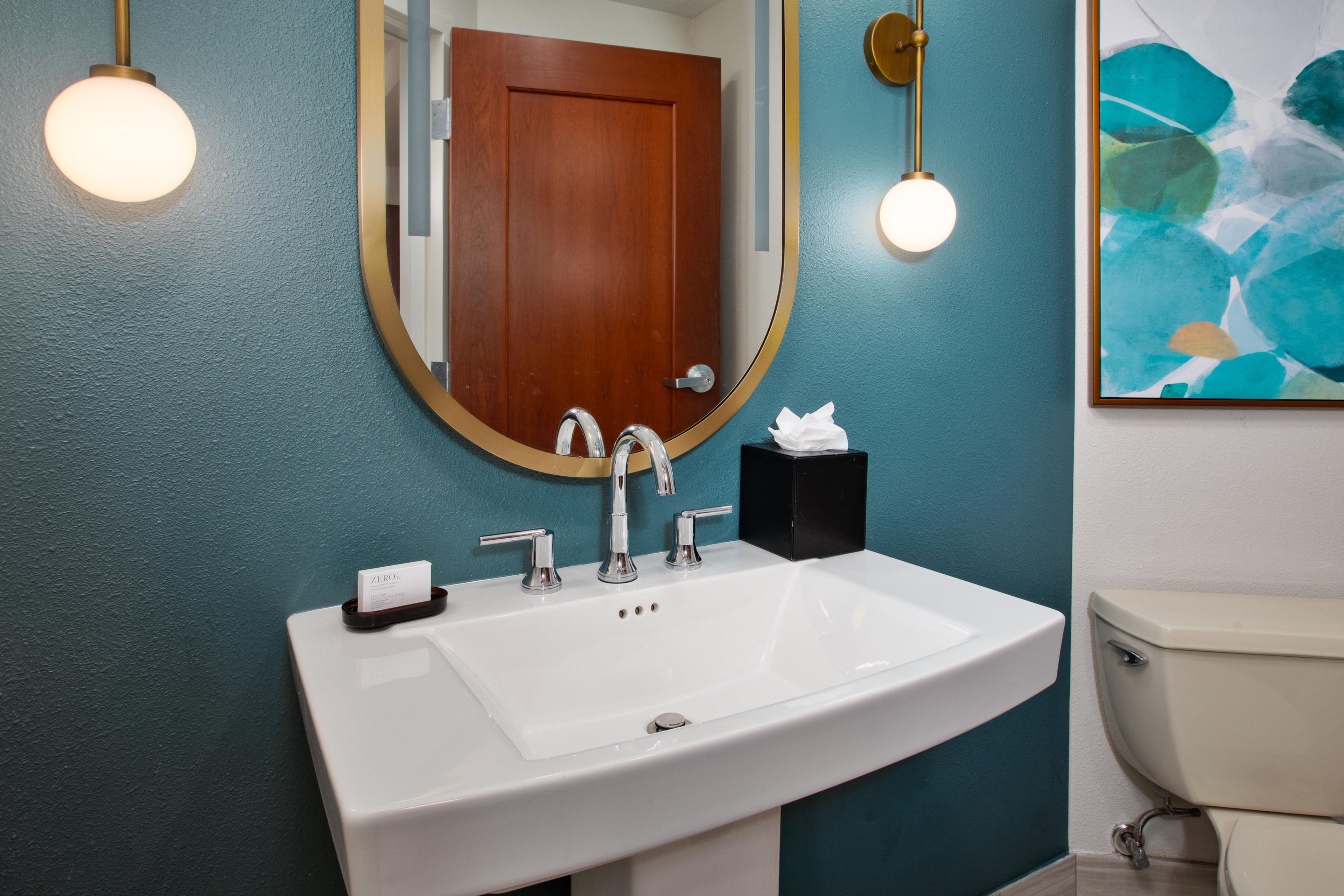 A bathroom with a white sink, two chrome faucets, a mirror with a gold frame, and a wooden door.
