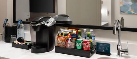 A hotel room with a coffee maker, a beverage dispenser, a snack bar, and a sink.