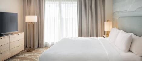 A hotel room with a large bed, a flat-screen TV, a desk with a lamp, and a view of the outdoors through the window.