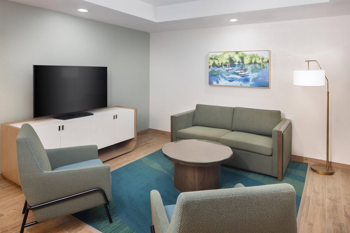 Een moderne woonkamer met een flatscreen televisie, een groene bank, twee fauteuils, een salontafel en een schilderij van een meer.