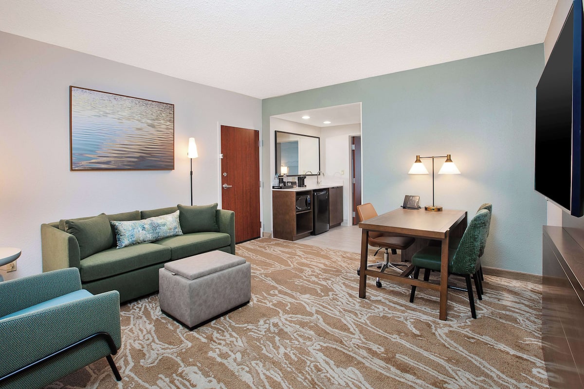 Een moderne hotelkamer met een groene bank, een eettafel met stoelen, een flatscreen-tv en een kitchenette.