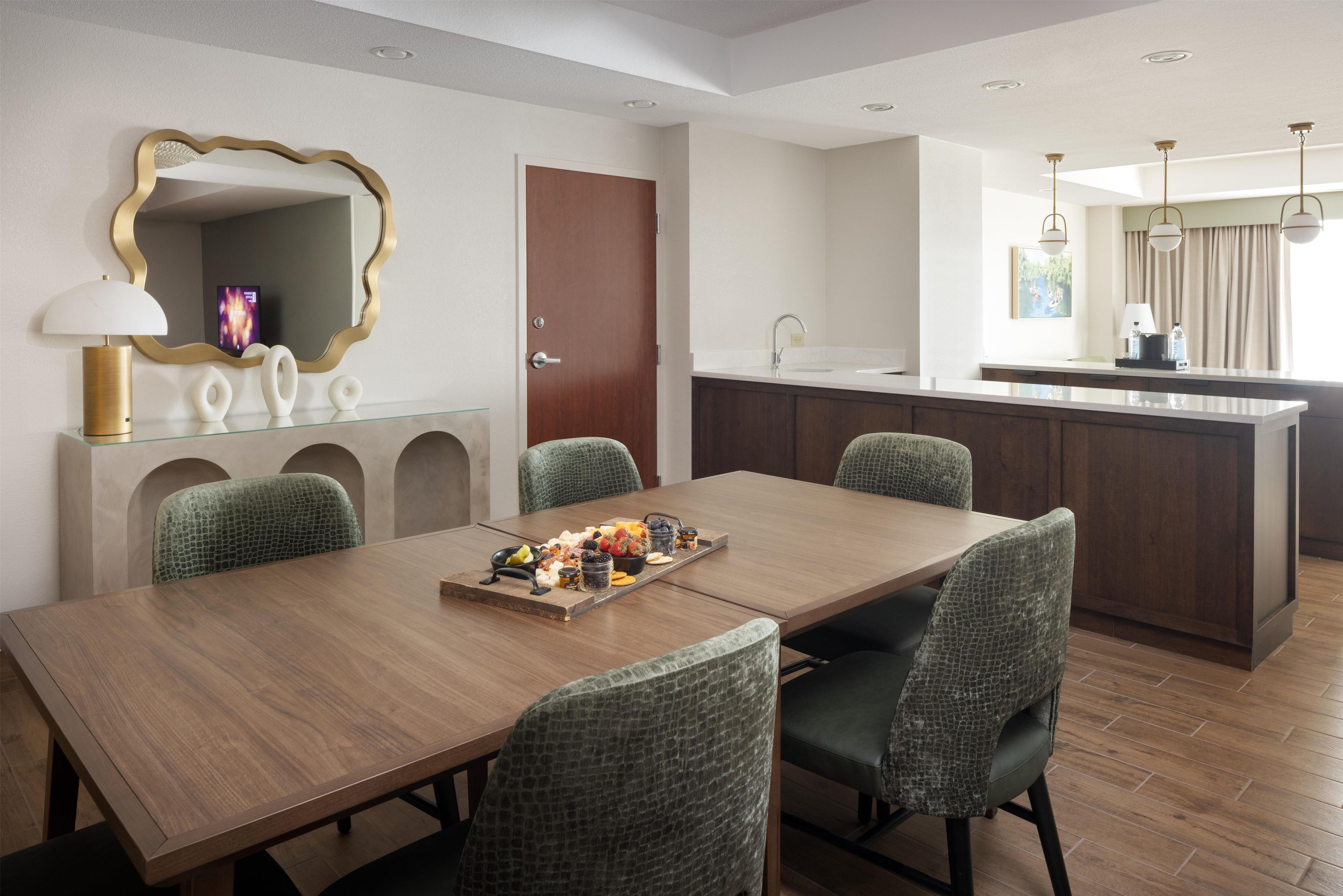 Ein moderner Hotel-Speisesaal mit einem Holztisch, grünen Sitzstühlen, einem Buffet mit verschiedenen Speisen, einem großen Spiegel und einer Küchenzeile mit Spüle und Schränken.