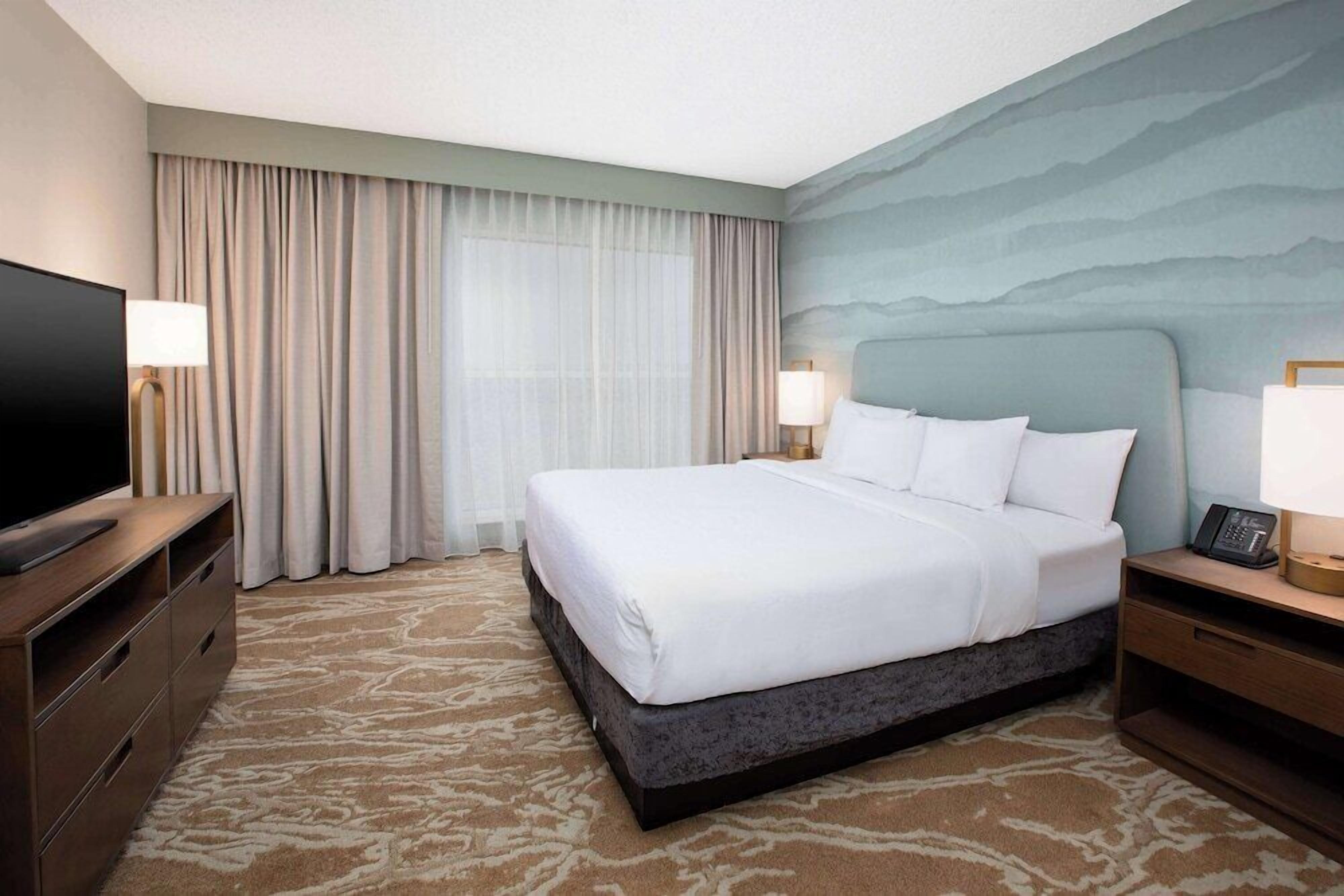 Quarto de hotel com uma cama grande, televisão de tela plana, escrivaninha com luminária e janela com cortinas.