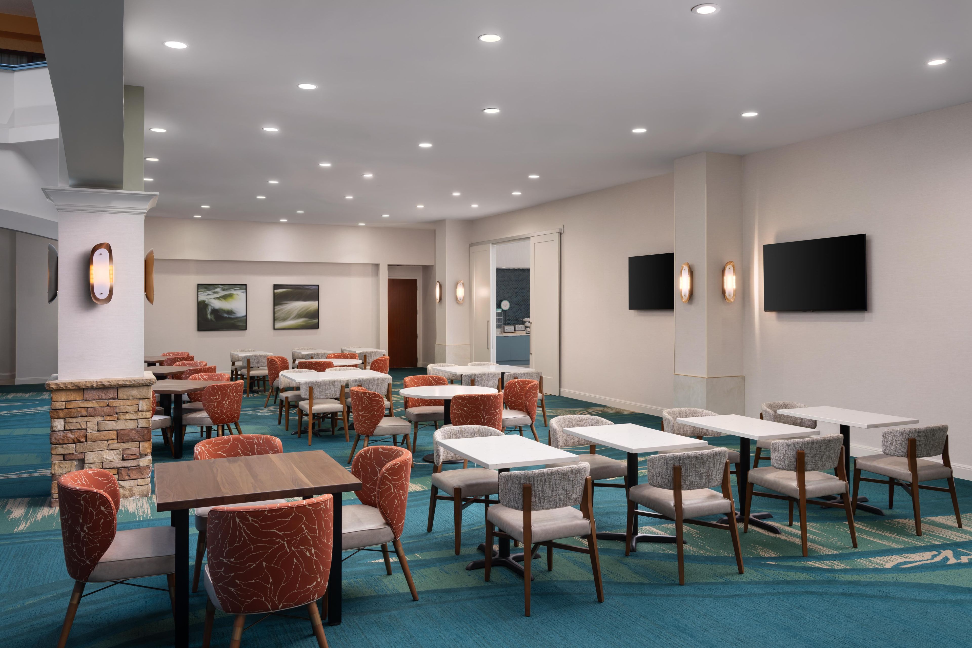 A hotel dining area with multiple tables and chairs, a stone accent wall, and a flat-screen TV mounted on the wall.
