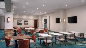 A hotel dining area with multiple tables and chairs, a stone accent wall, and a flat-screen TV mounted on the wall.