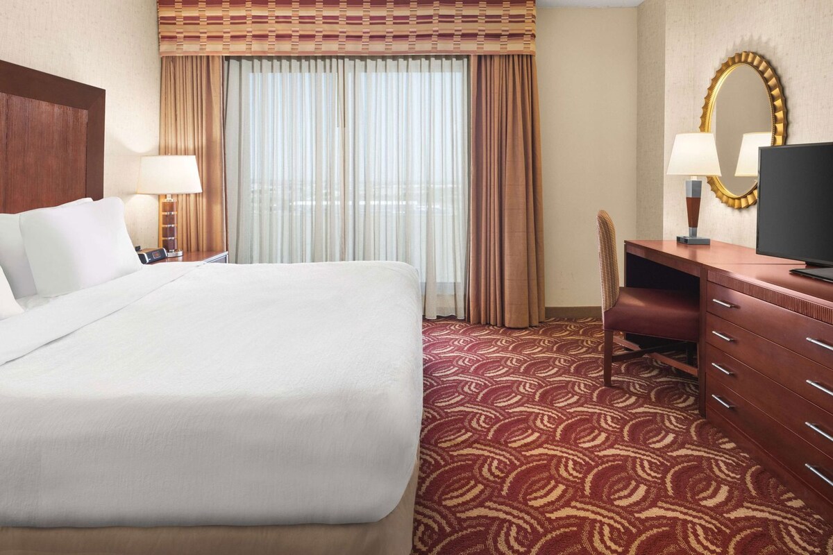Una habitación de hotel con una cama grande, un escritorio con televisor, una silla y un espejo.