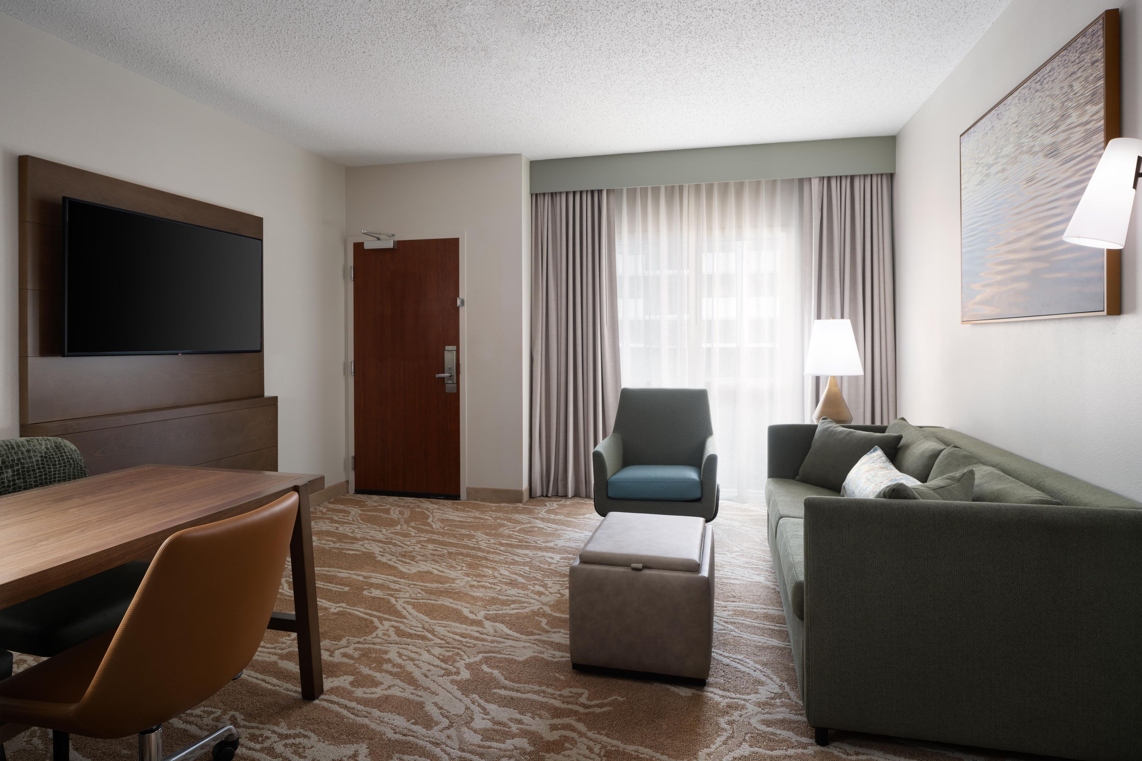 A hotel room with a flat-screen TV, a wooden dining table, a sofa, a chair, and a lamp.