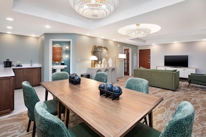 Habitación de hotel con una mesa de comedor, sillas, un sofá, televisión y un baño con lavamanos y espejo.