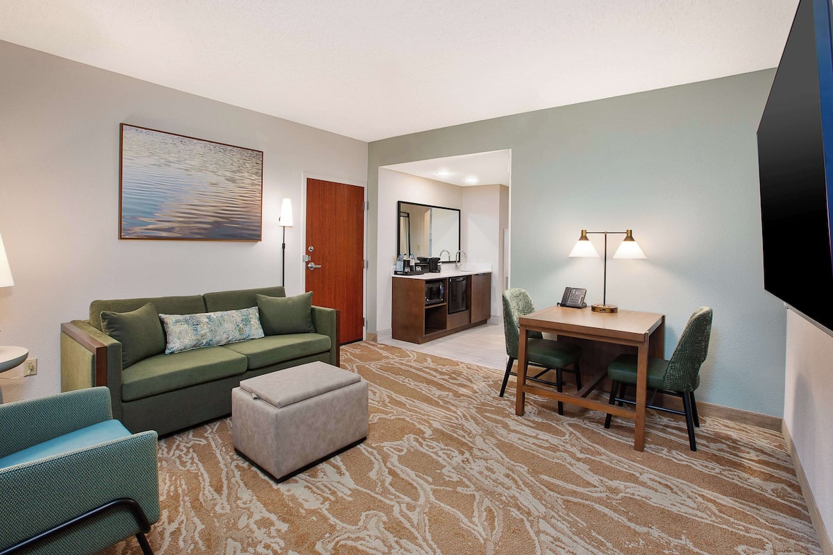 Een hotelkamer met een groene bank, een eettafel met stoelen, een flatscreen televisie, een schilderij en een keukenhoek.
