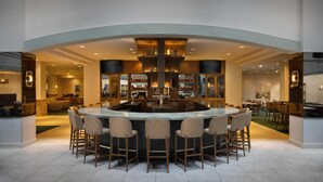 Uma área de bar moderna com ilha central e banquetas, rodeada por assentos e teto alto com iluminação embutida.
