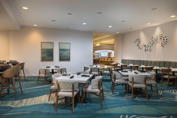 Un comedor de hotel con mesas y sillas redondas, una alfombra estampada y una pared con arte abstracto.