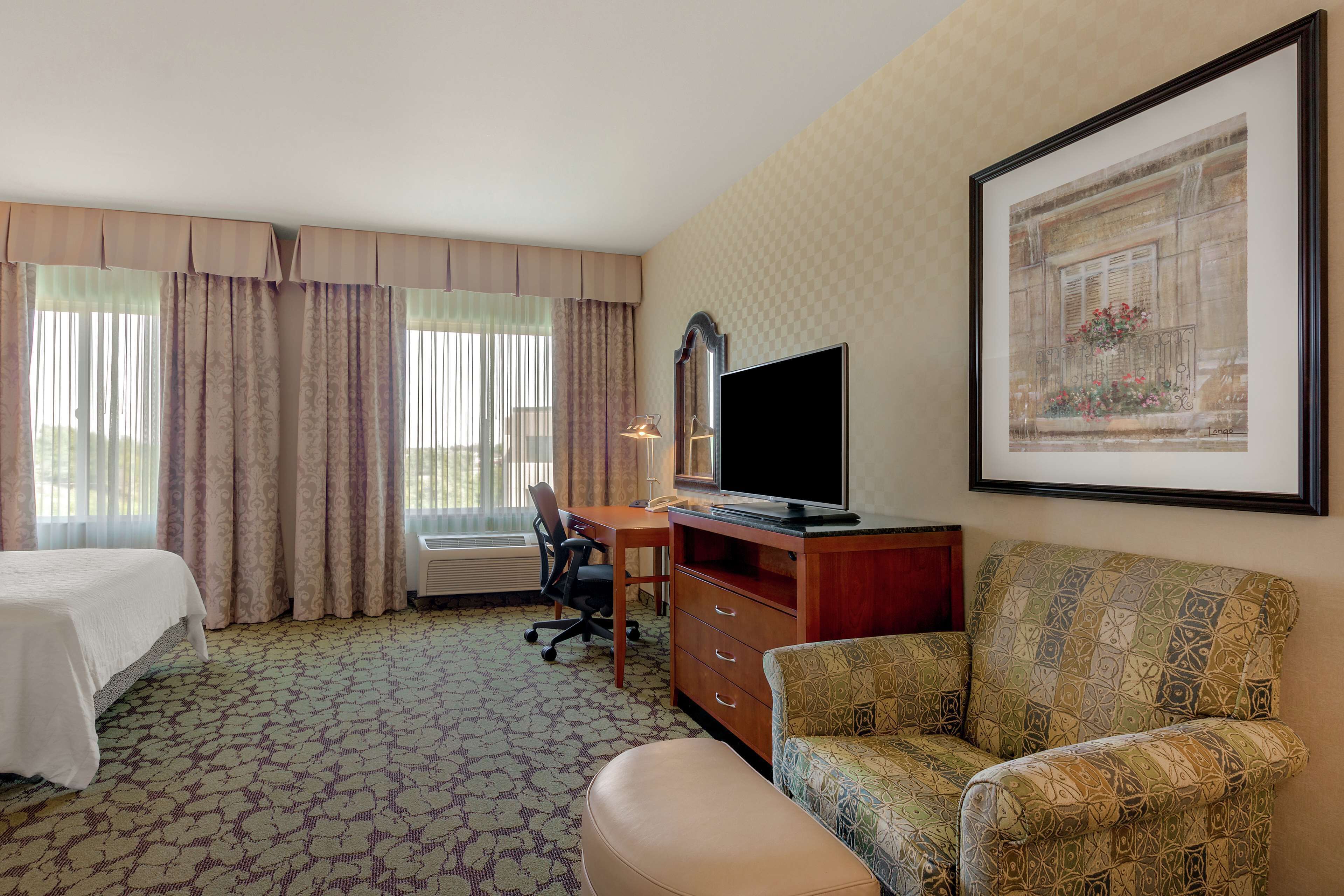 A hotel room with a bed, a desk with a TV, a chair, a sofa, and a window with curtains.