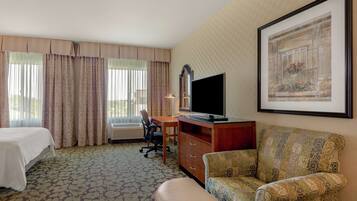 A hotel room with a bed, a desk with a TV, a chair, a sofa, and a window with curtains.