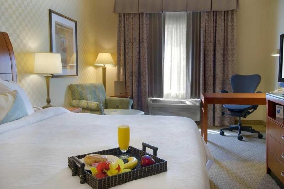 A hotel room with a bed, a desk, a chair, a lamp, a window with curtains, and a tray with fruits and juice.