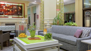 A living room with a grey sofa, a white coffee table, and a green plant. There is a glass jar with lemons and a glass jar with green apples on the table. - Hilton Garden Inn Sacramento Elk Grove (Elk Grove)