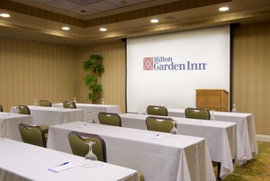 Un salón de conferencias con mesas y sillas, una pantalla de proyector que muestra el logotipo de Hilton Garden Inn y un atril.