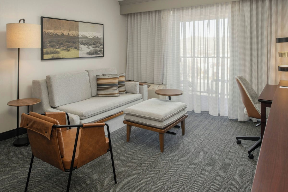 A modern hotel room with a sofa, ottoman, brown leather chair, and a desk with a lamp.
