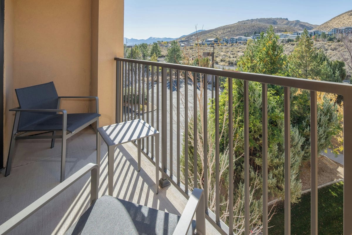 A balcony with a metal railing, a blue chair, and a view of a hilly landscape with trees and buildings in the distance.