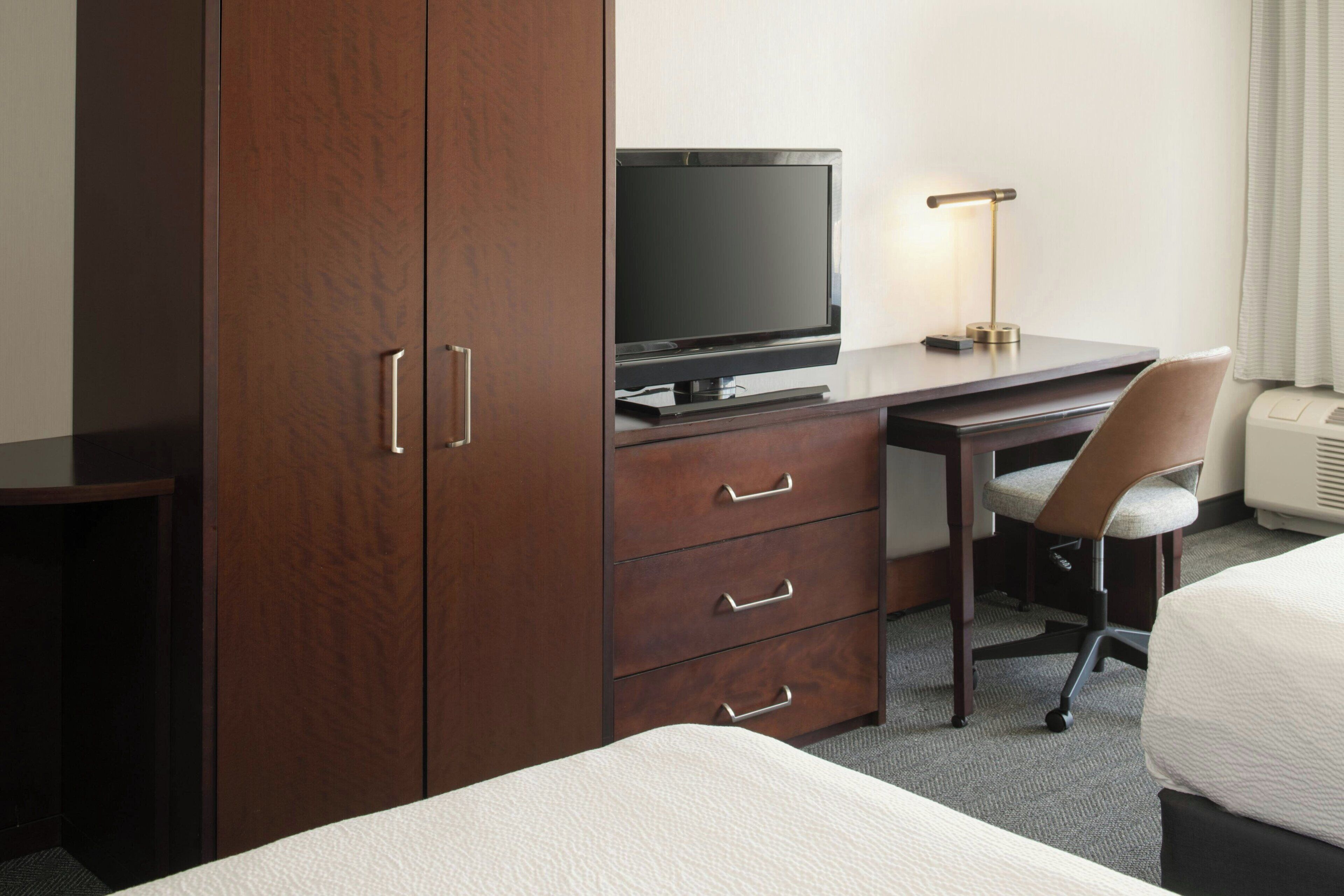 A hotel room with a wooden wardrobe, a desk with a television, a chair, and a bed.