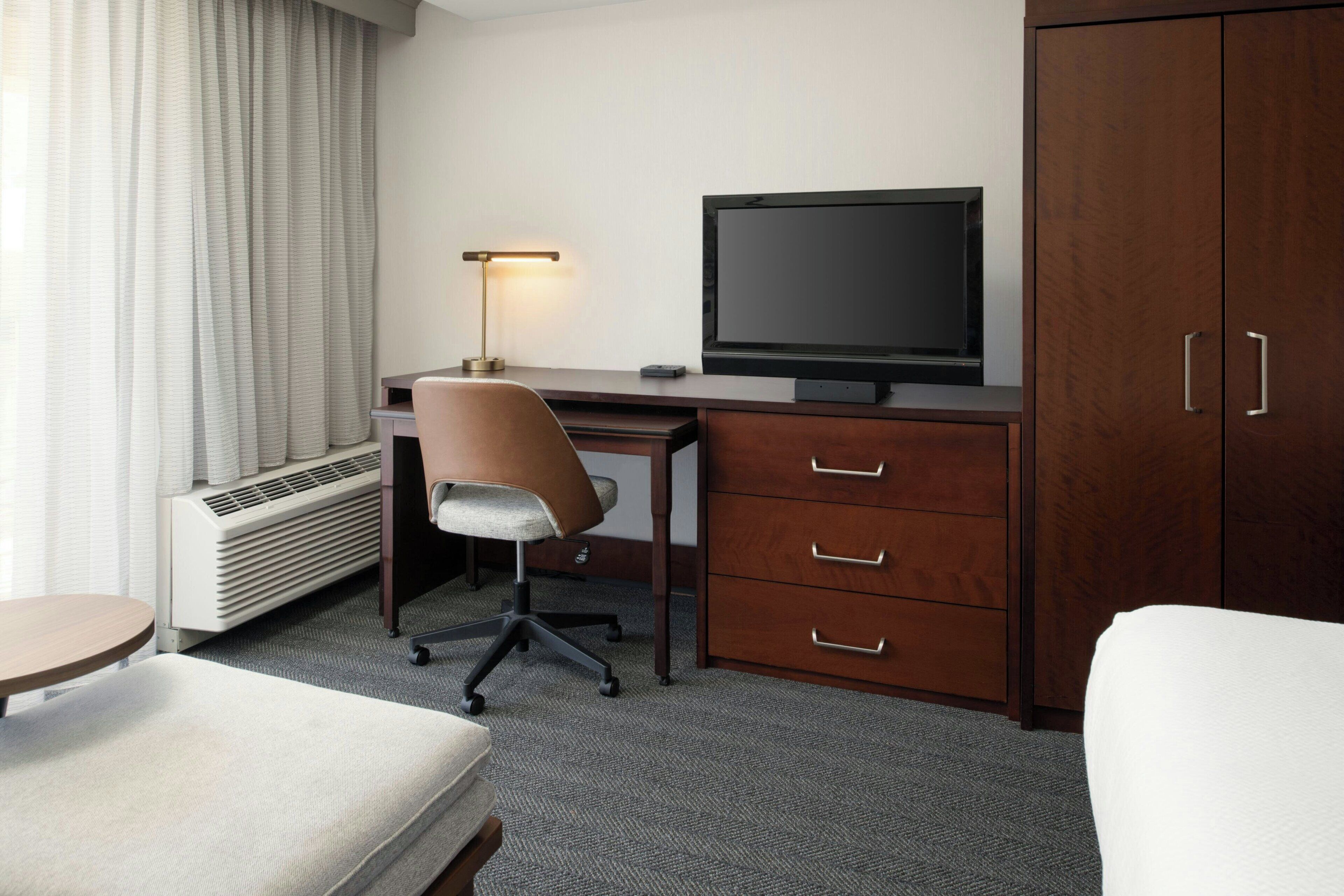 Quarto de hotel com secretária, cadeira, televisão, guarda-roupa e cama.