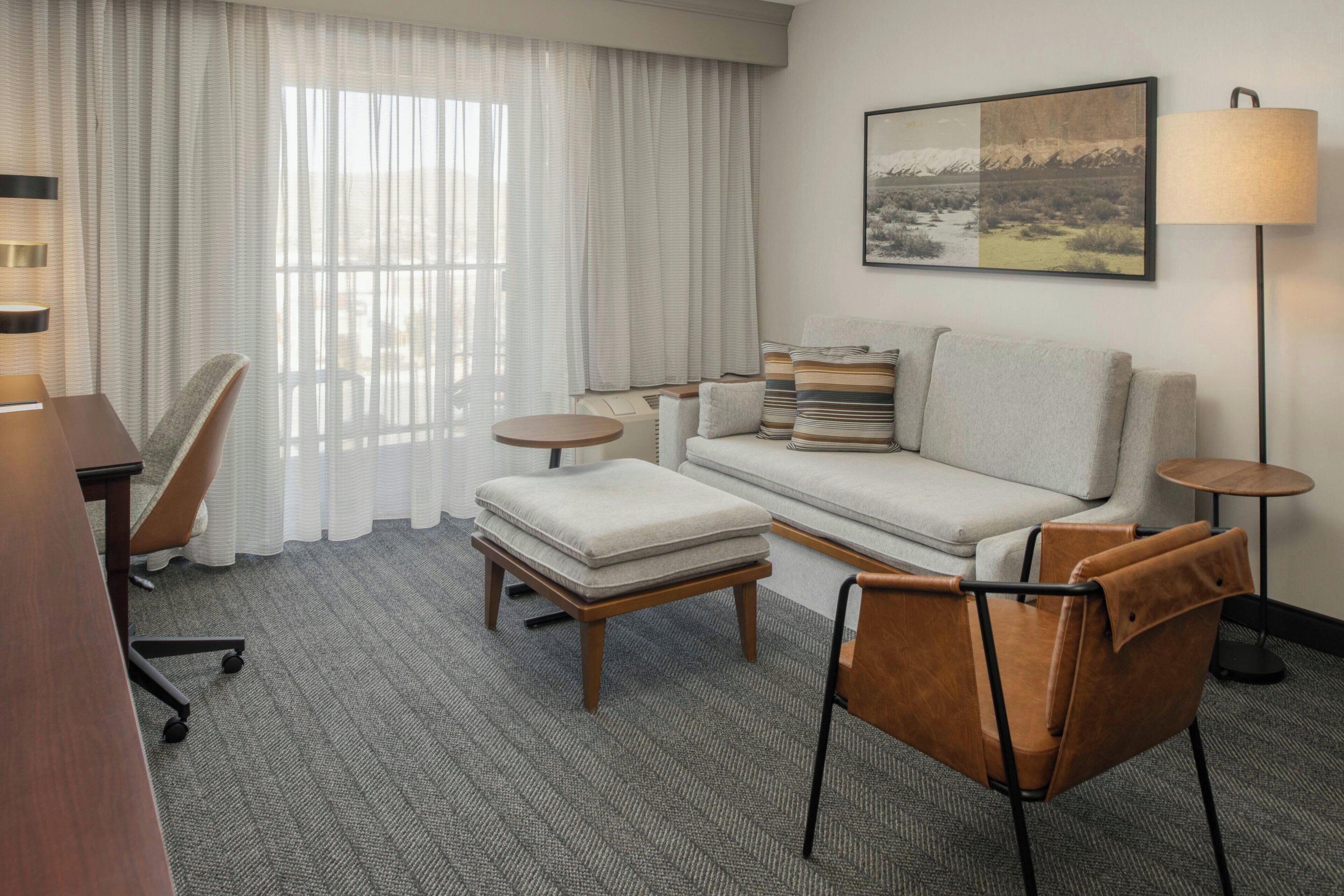 A hotel room with a sofa, armchair, ottoman, and a desk with a chair.