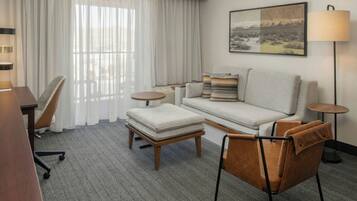 A hotel room with a sofa, armchair, ottoman, and a desk with a chair.