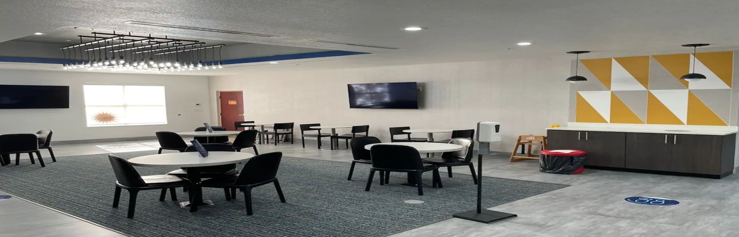 En moderne hotellobby med runde borde og stole, et fladskærms-TV, et tekøkken og et geometrisk vægdesign.