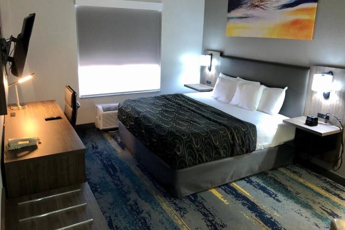 Ein Hotelzimmer mit Bett, Nachttischen, Schreibtisch, Stuhl, Fenster mit Jalousien und einem Bild an der Wand.