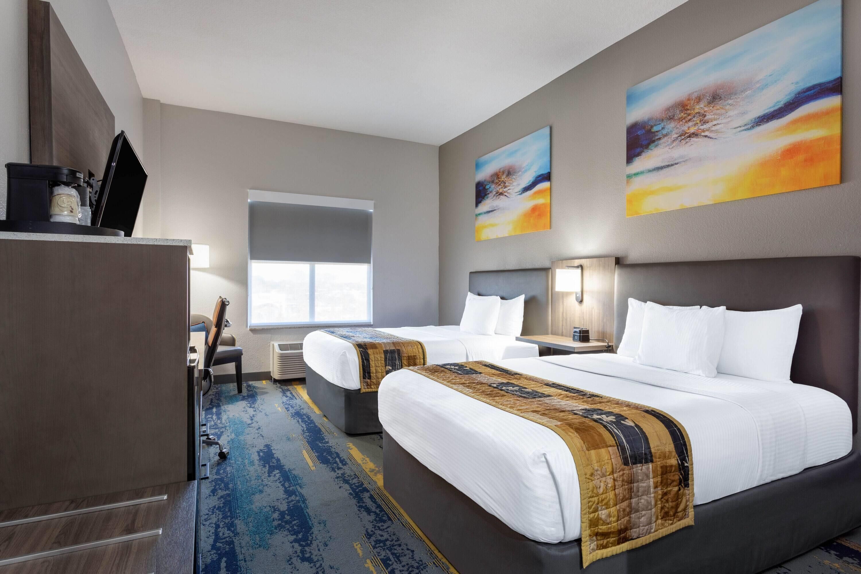 Ein Hotelzimmer mit zwei Betten, einem Schreibtisch, einem Stuhl, einem Fenster und abstrakten Bildern an der Wand.