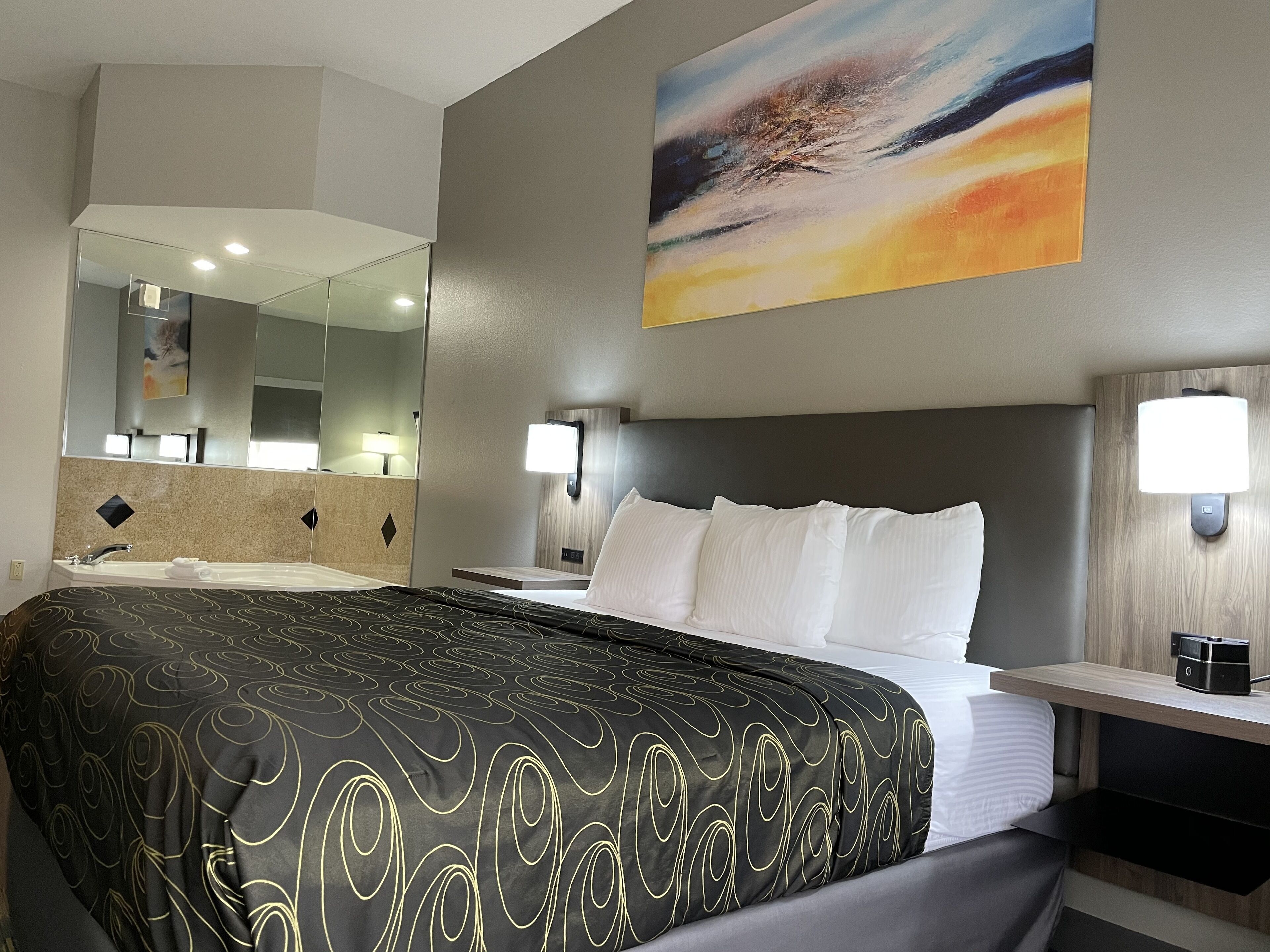 A hotel room with a large bed, a bathroom with a mirror, and a painting on the wall.
