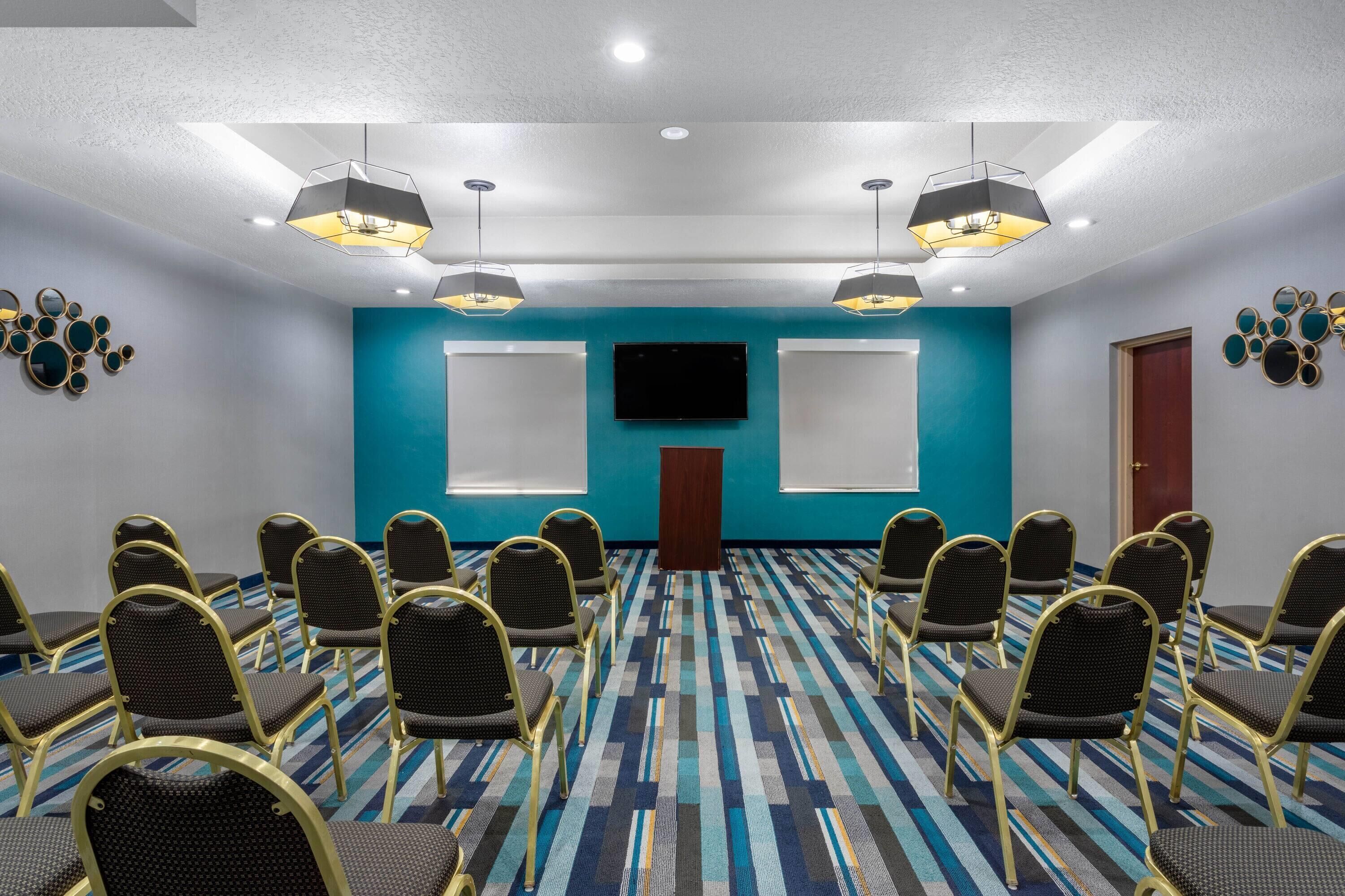 A conference room with a blue accent wall, a flat-screen TV, and a podium. Rows of chairs are arranged facing the TV.