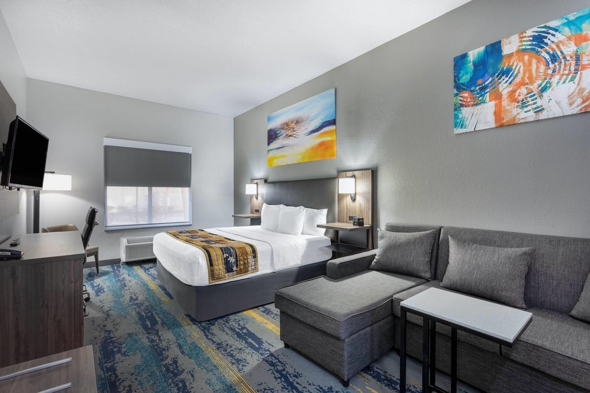 Ein Hotelzimmer mit Bett, Sofa, Schreibtisch, Fernseher und einem Gemälde an der Wand.