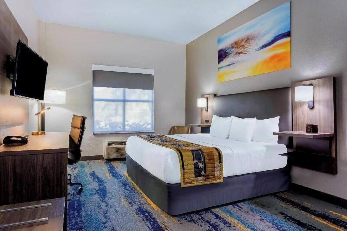 Ein Hotelzimmer mit Bett, Schreibtisch, Stuhl, Fernseher, Kunstwerken und einem Fenster.