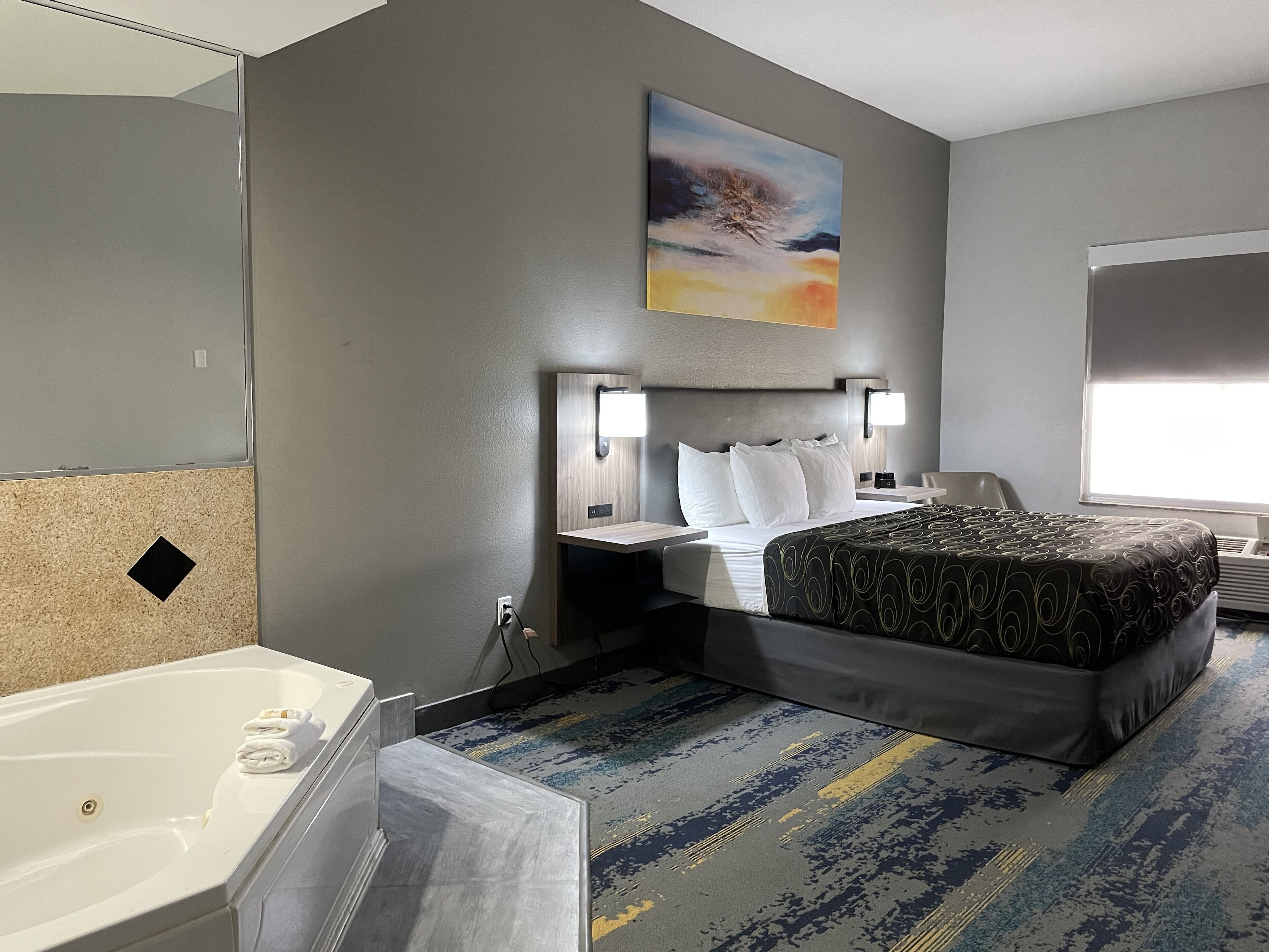 Ein Hotelzimmer mit Bett, Nachttischen, Lampe, einem Bild, einem Fenster und einem Badezimmer mit Badewanne und Handtuchhalter.