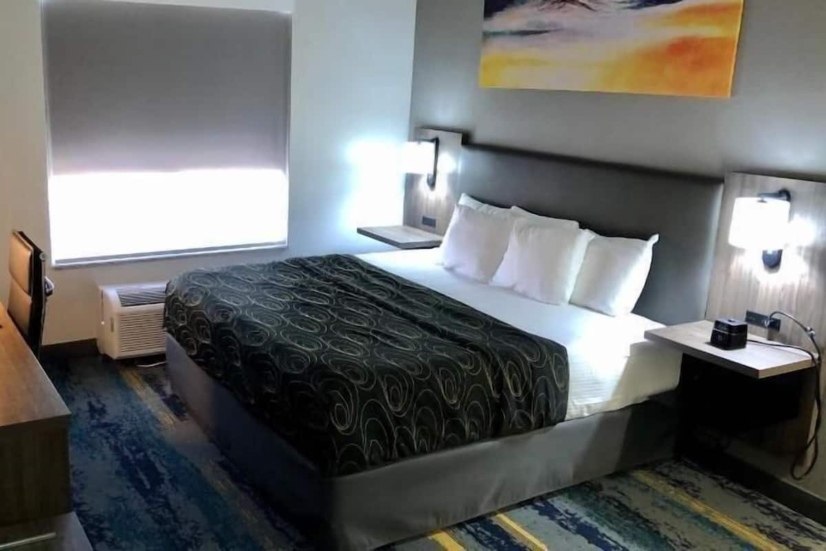 A hotel room with a bed, bedside tables, a desk, a chair, a TV, a window, and a painting on the wall.