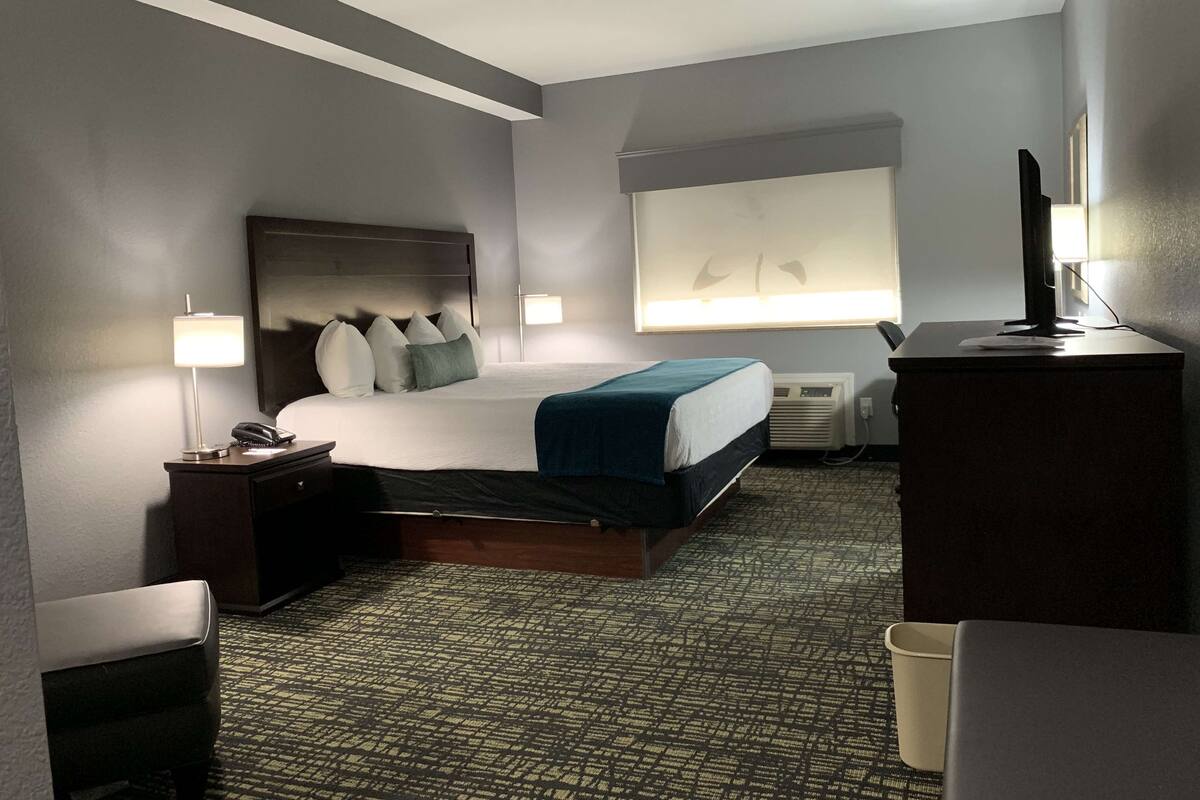 Une chambre d’hôtel avec un grand lit, des lampes de chevet, un bureau avec un ordinateur et une fenêtre avec des stores.