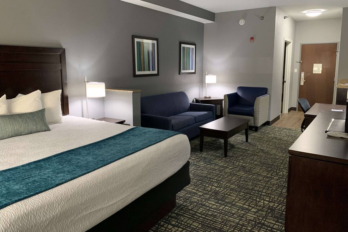 Une chambre d’hôtel avec un grand lit, un canapé, un bureau et une chaise. Il y a deux tableaux encadrés au mur et une porte sur la droite.