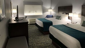Quarto de hotel com duas camas, uma mesa, uma cadeira, televisão e janela com persianas.