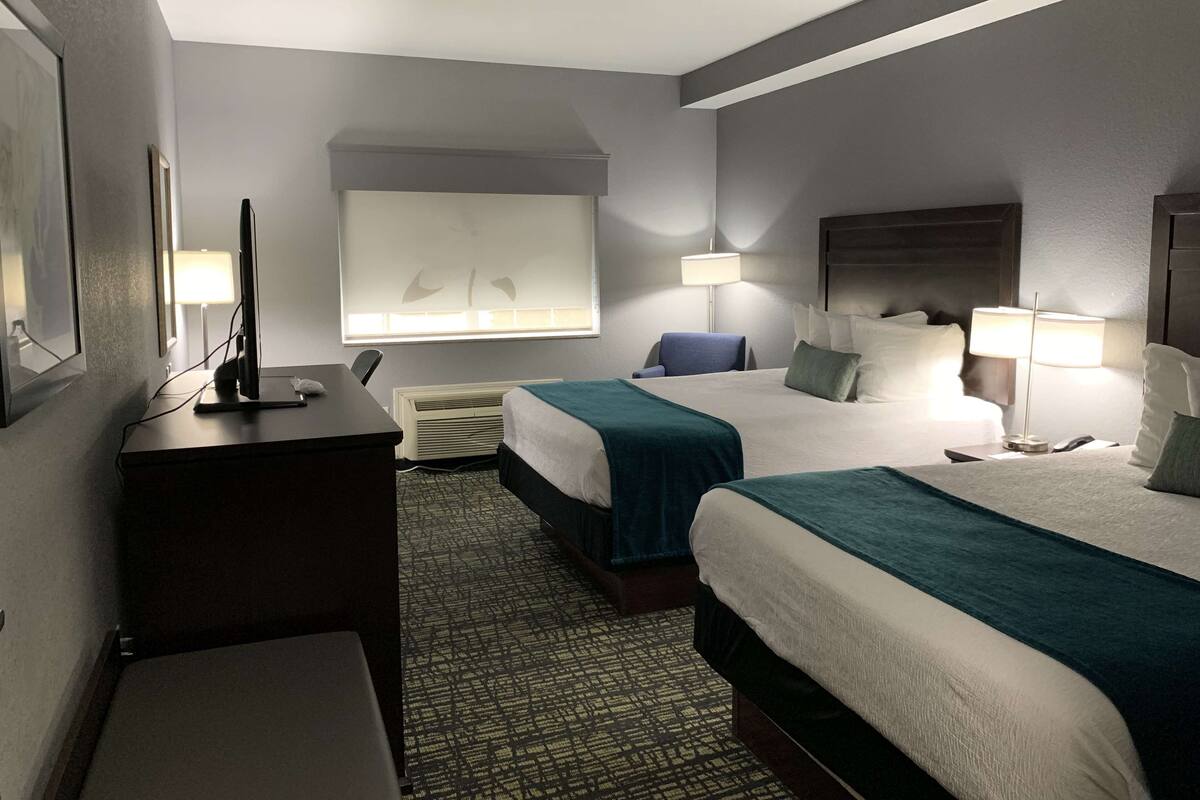 Une chambre d’hôtel avec deux lits, un bureau, une chaise, une télévision et une fenêtre avec des stores.