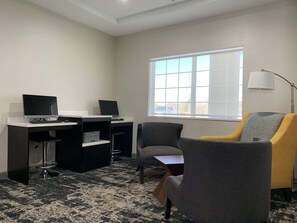 Un espacio de oficina moderno con dos escritorios, uno con computadora y teclado, y otro con una impresora. Hay un sillón gris cómodo y un sillón amarillo con una lámpara.