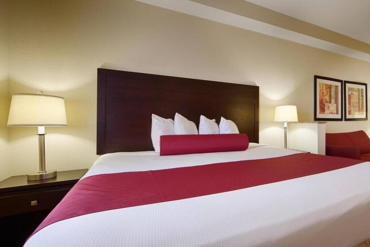 Une chambre d’hôtel avec un grand lit, deux lampes de chevet, un coussin décoratif rouge et des œuvres d’art encadrées accrochées au mur.