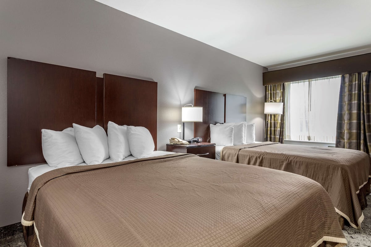 A hotel room with two beds, a nightstand, a lamp, and a window with curtains.