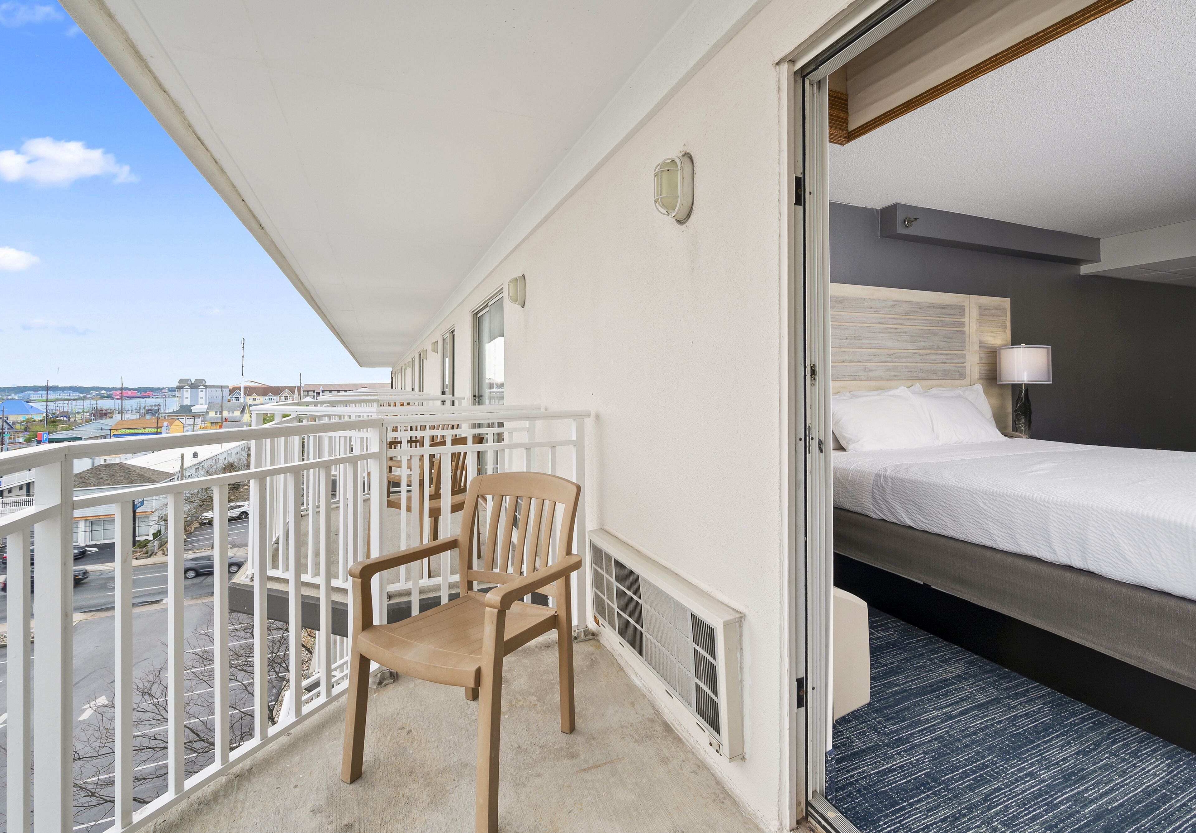 A balcony with a wooden chair, a bed with white bedding, and a view of the city.